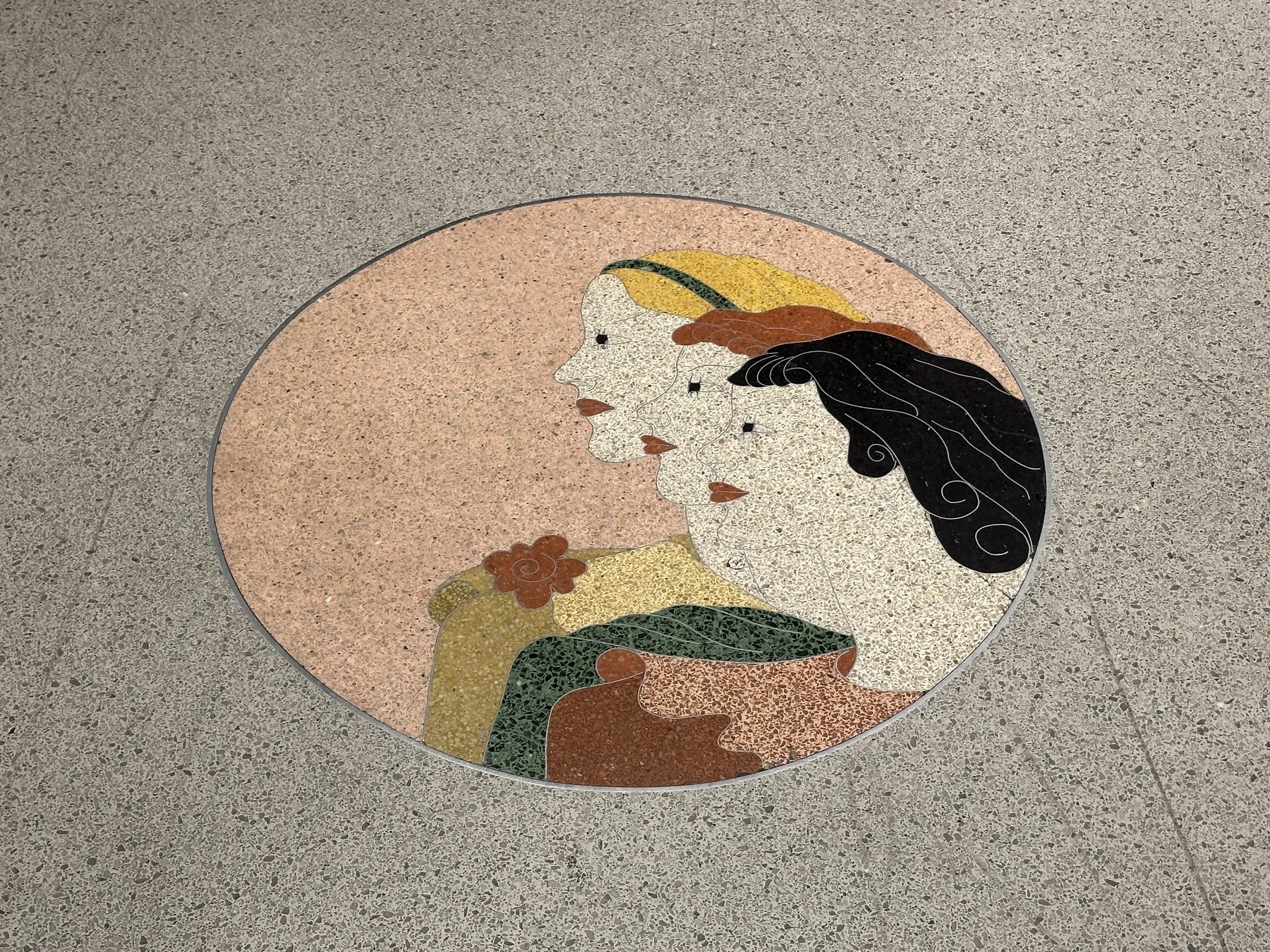 An oval terrazzo medallion set into a grey terrazzo shop floor showing three women's profiles in coloured marble chips outlined by brass divider strips