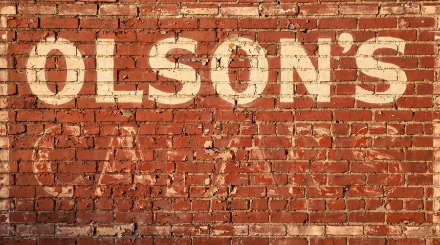 Ghost sign palimpsest — a faded painted advertisement overlapping the illegible remains of an older ad on a red brick wall
